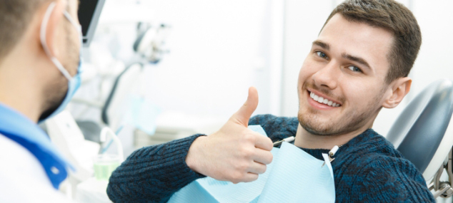 A young man sitting in a dental chair smiles confidently and gives a thumbs-up to the dentist, who is partially visible and wearing a face mask.